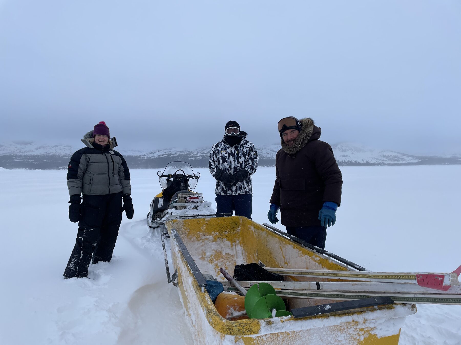 Three people in heavy winter parkas standing beside a snowmobile and yellow sled loaded with field equipment on the ice.