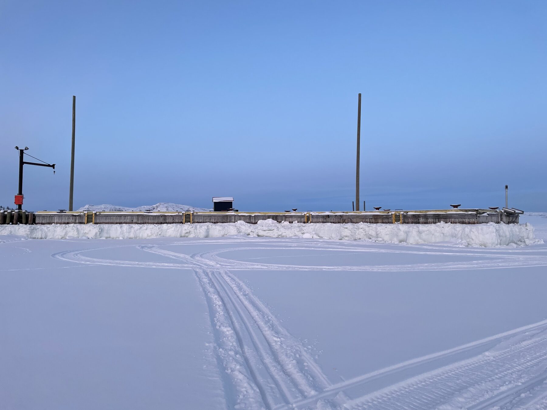 Snowmobile tracks crossing sea ice in front of a wooden wharf near Nain at dusk.