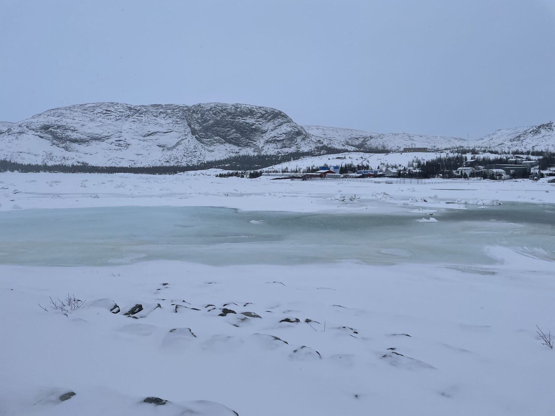 Coastal mountain and partially frozen water near a small Arctic community under overcast skies.