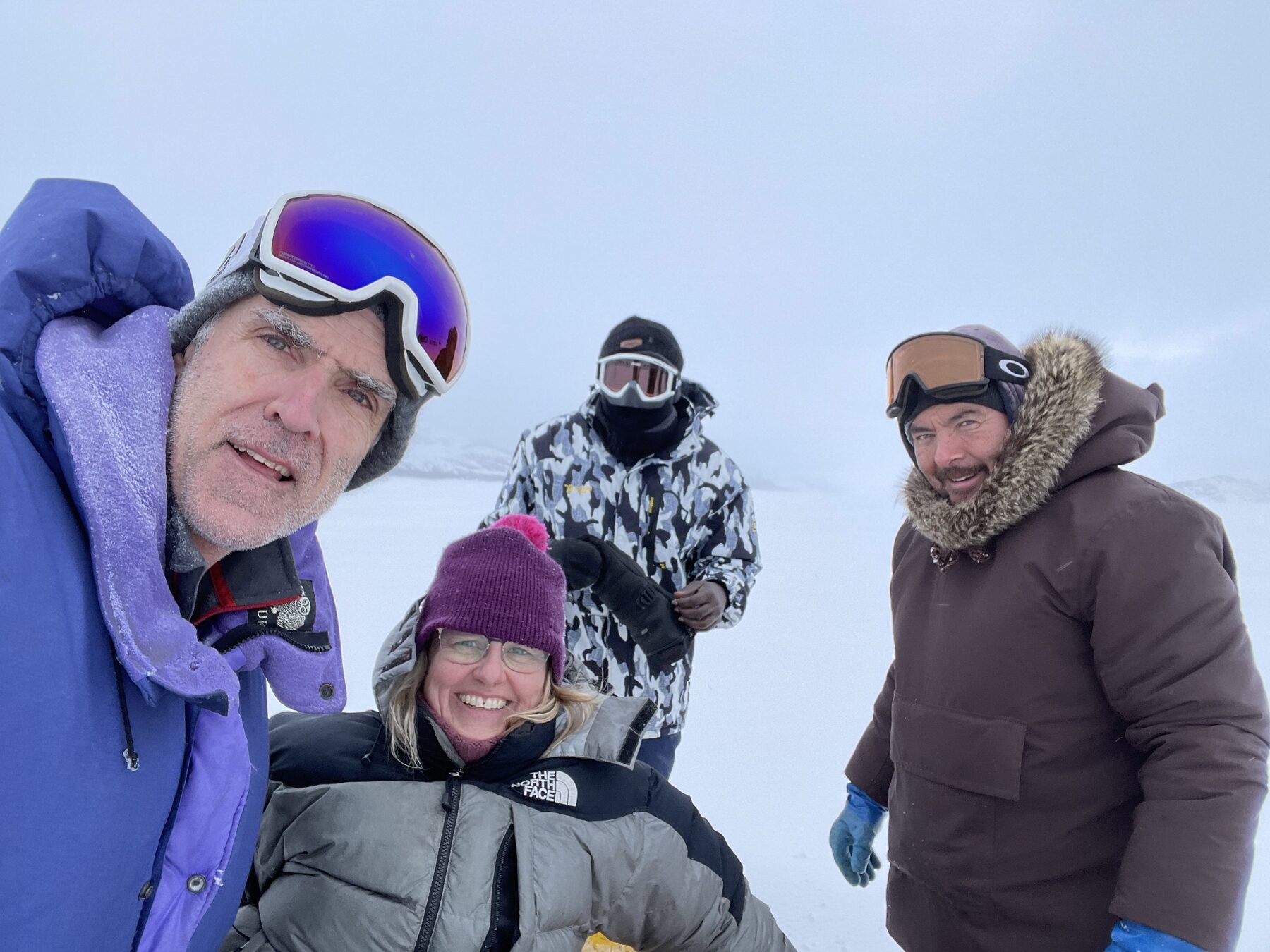 Group of researchers in parkas and ski goggles gathered close together on a snowy plain under grey skies.