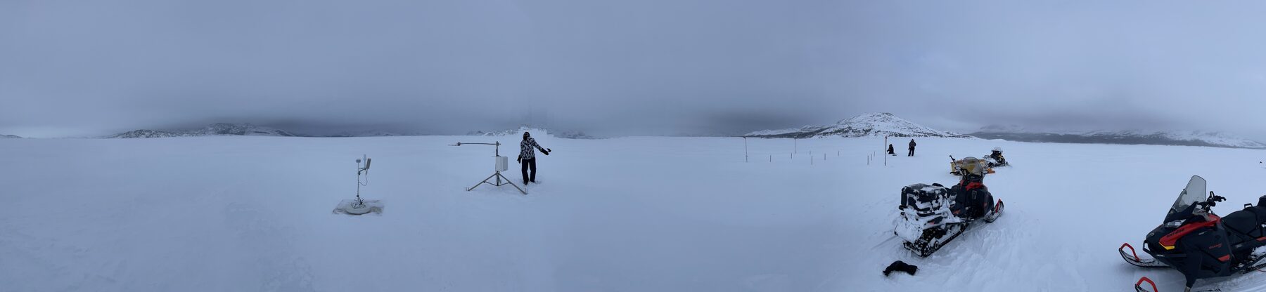 Wide panorama of a snow-covered field with an instrument tripod, a researcher, and snowmobiles under low cloud.